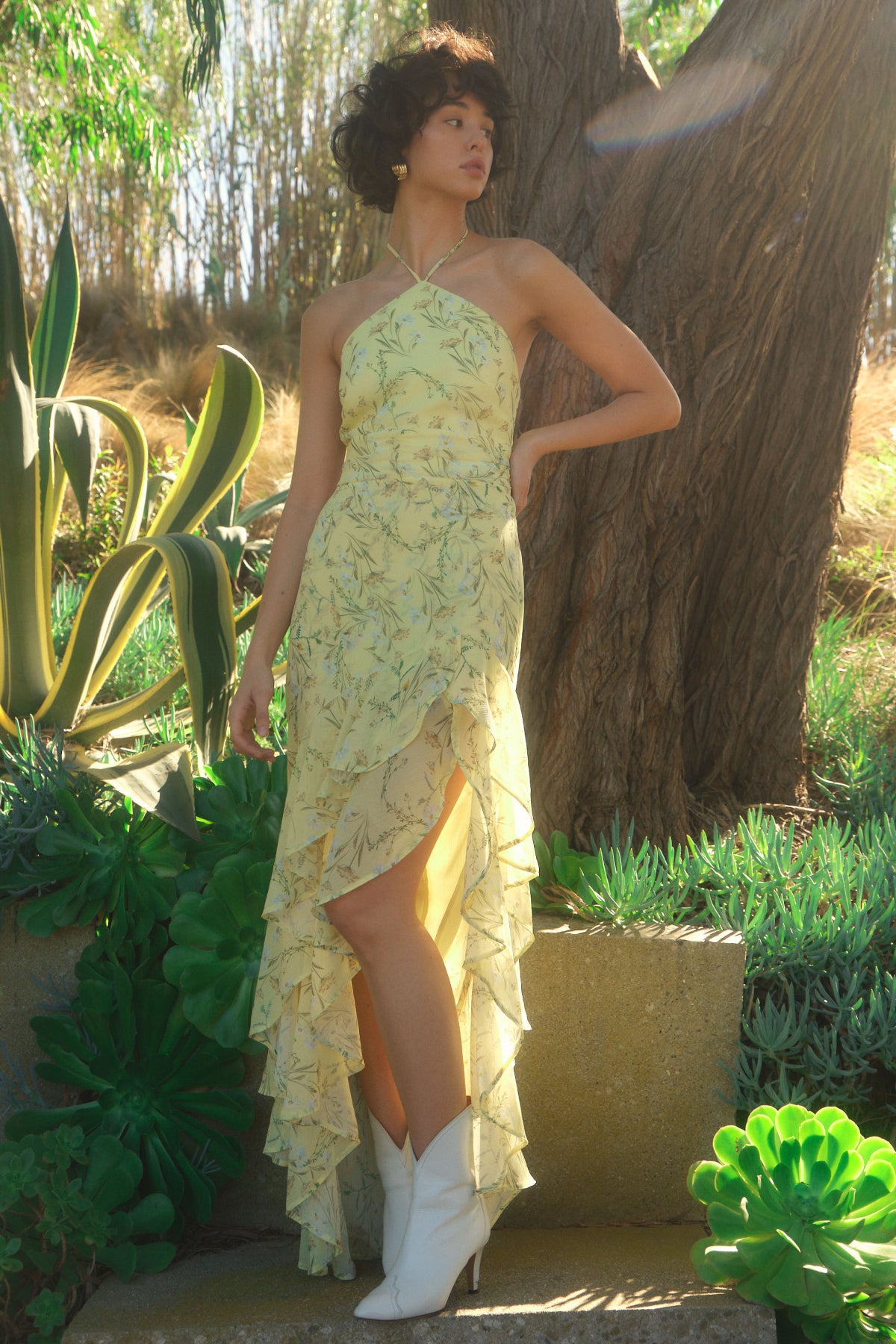 Woman in a yellow floral dress and white boots standing in a garden setting.