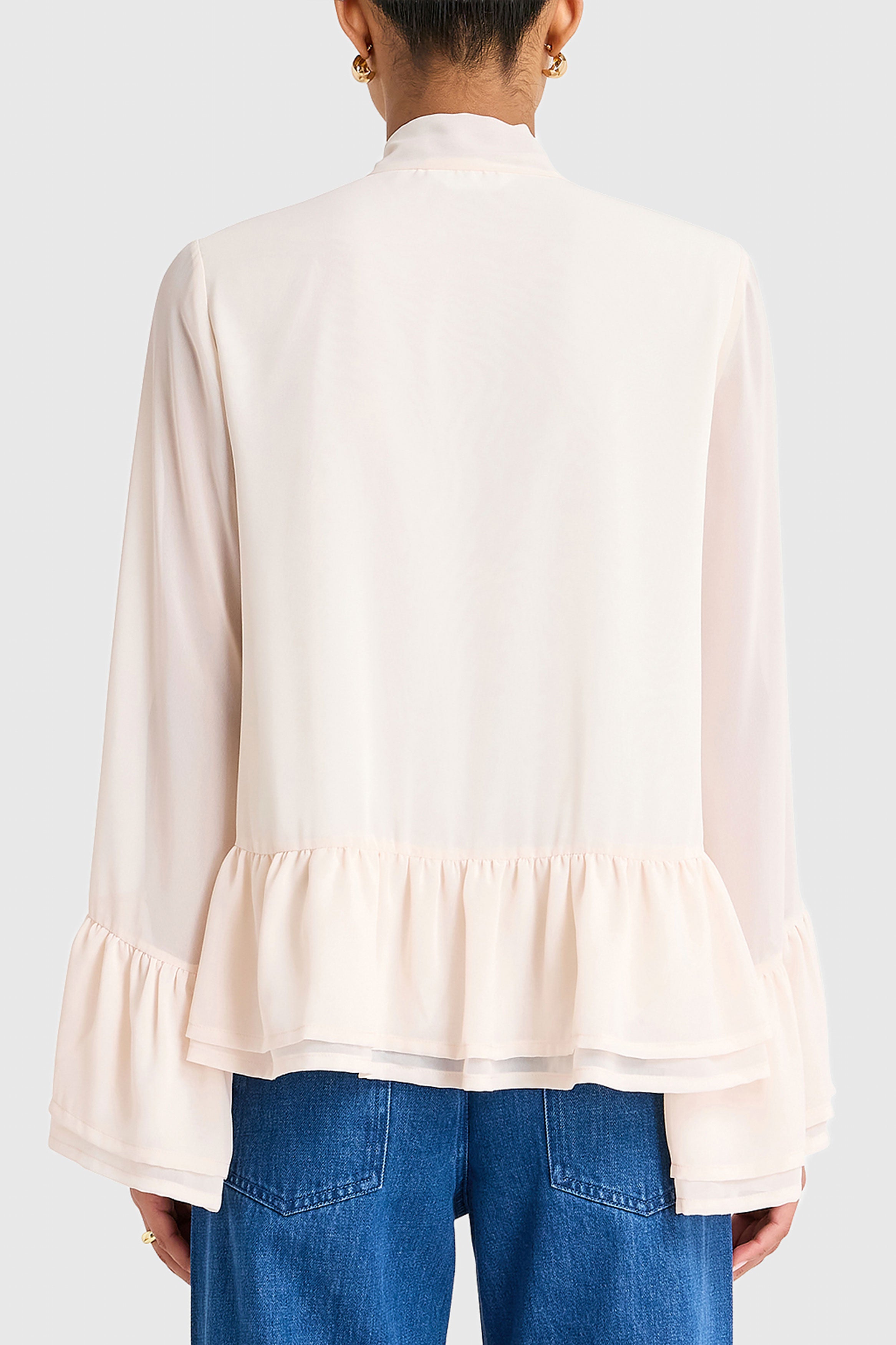 Person wearing a light pink blouse with ruffled layers and blue jeans on a white background
