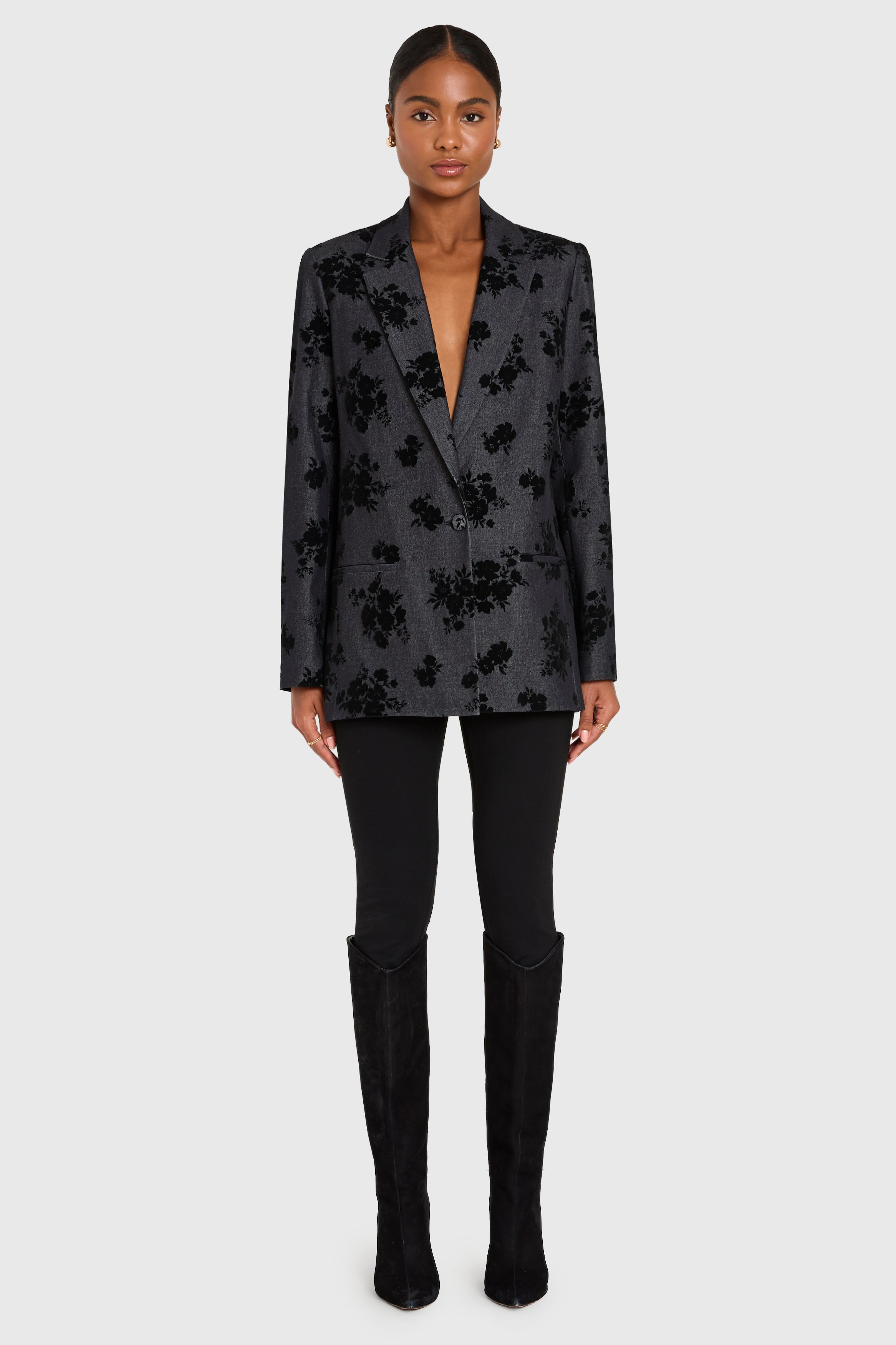 Woman wearing a dark floral blazer, black pants, and black knee-high boots on a white background