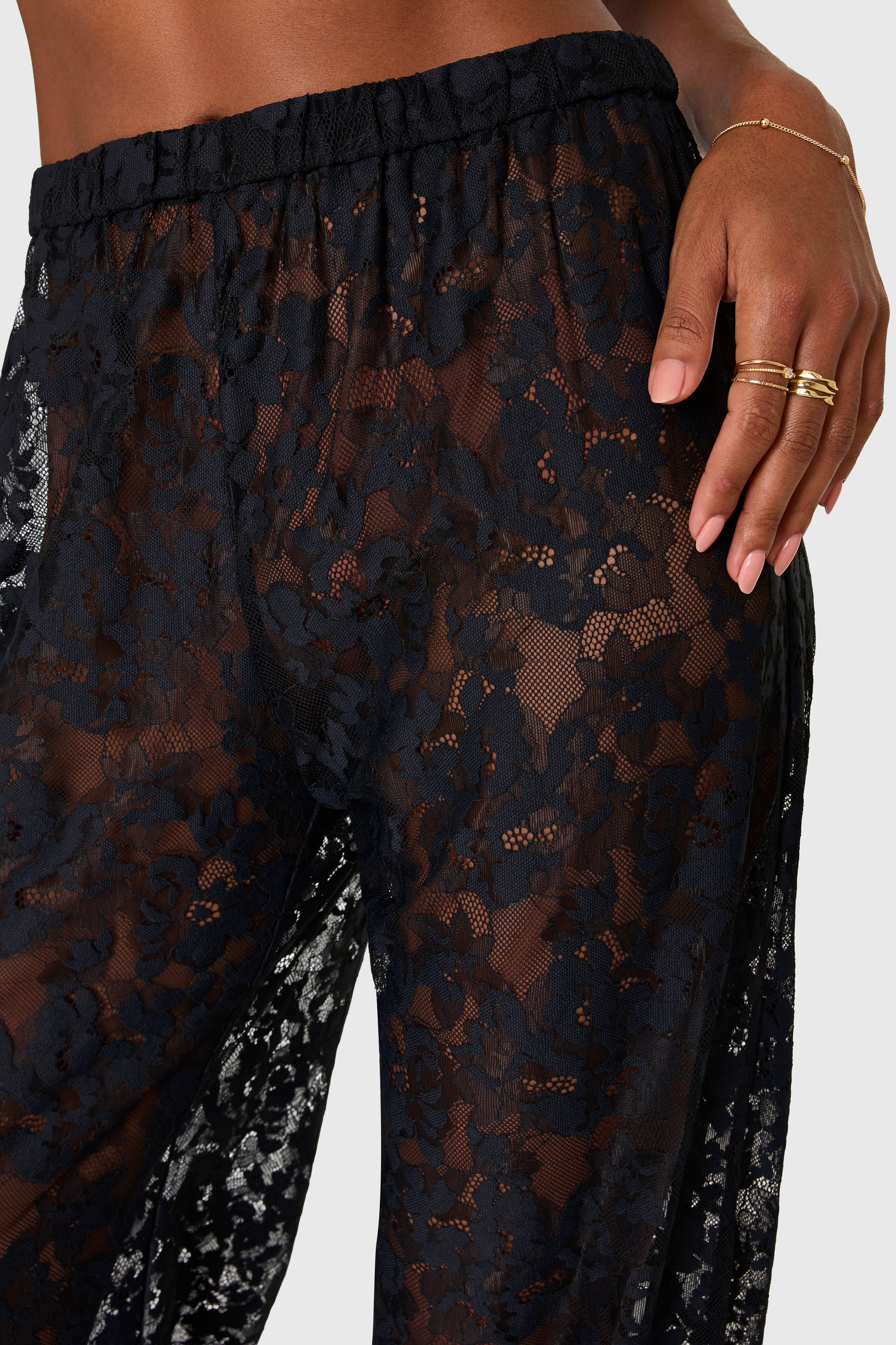 Black lace pants with a close-up of a hand wearing gold rings on a white background