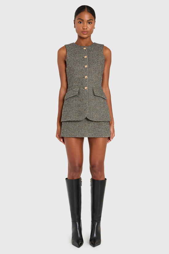 Woman wearing a sleeveless tweed dress with buttons and black knee-high boots on a white background view 1
